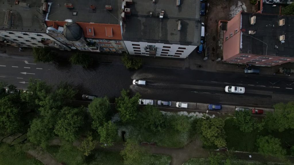Cars navigate through a flooded street in an urban area, captured from above.